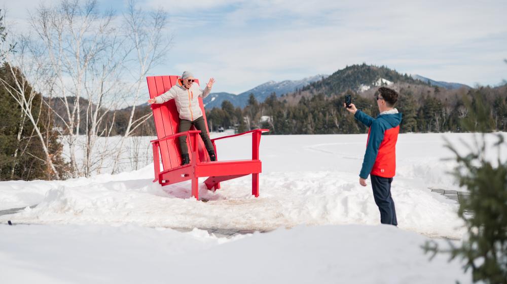 A man poses for a photo on an oversized Adirondack chair on a snowy lawn.