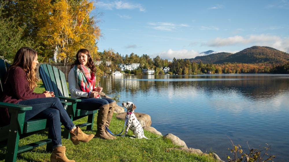 Two women and a dog sit in Adirondack chairs by a calm lake surrounded by fall foliage.