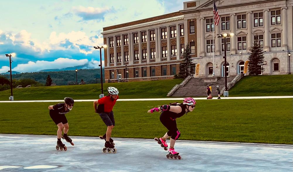 Three adults in helmets and skating clothes inline skate in a row, with other skaters and a school in the background.